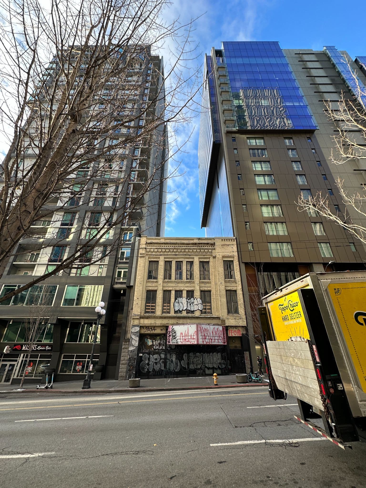 The Seven Seas Building today, seen from across 1st Avenue: the tan-brick three-story facade boarded and tagged, wedged between two modern towers